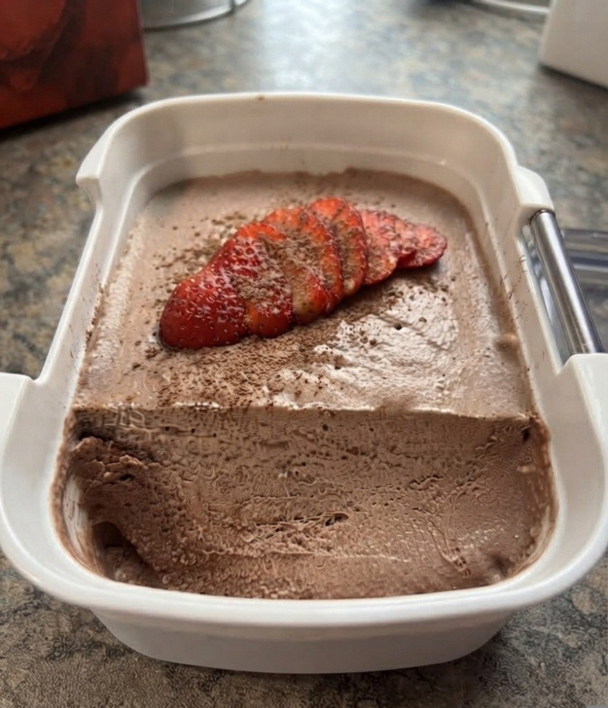 A container of chocolate ice cream topped with sliced strawberries and a dusting of cocoa powder, placed on a countertop.