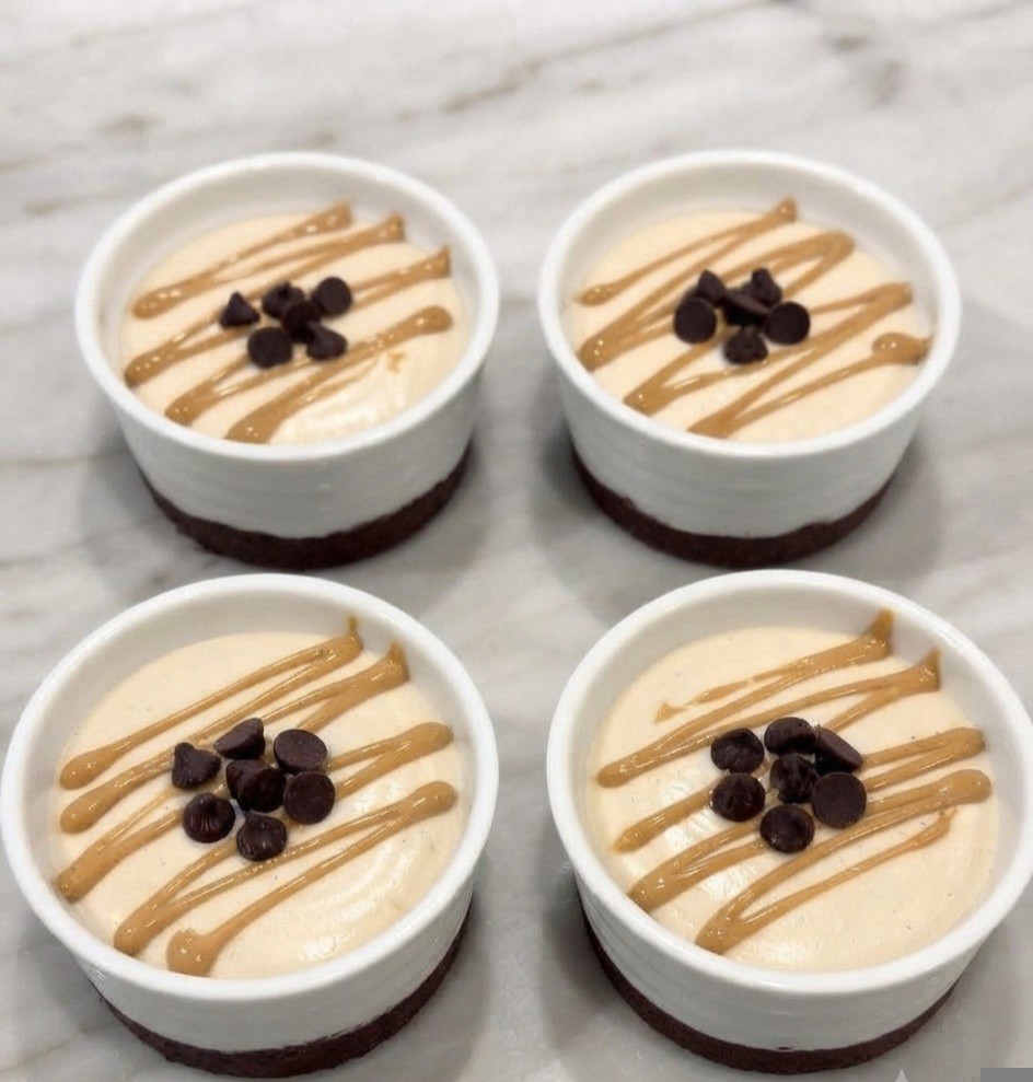 Four small cups of dessert with a beige creamy topping, drizzled with peanut butter and topped with chocolate chips, placed on a marble surface.