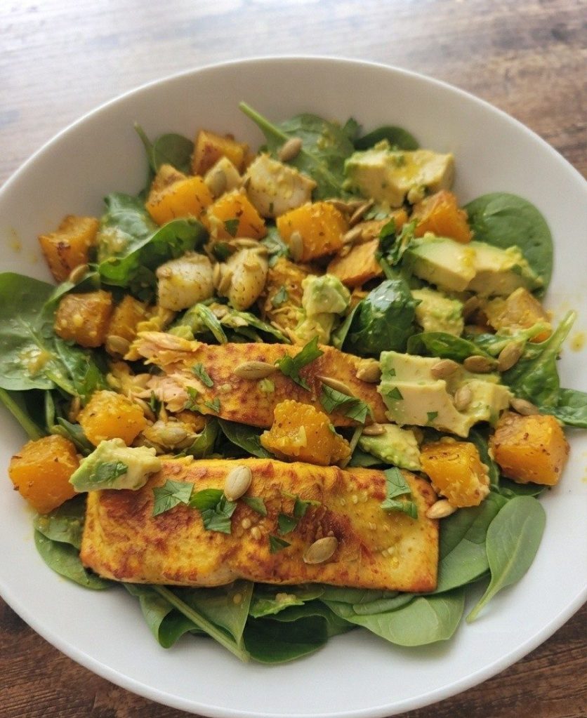 A fresh salad featuring spinach, roasted butternut squash, avocado chunks, and grilled chicken, topped with pumpkin seeds and herbs.