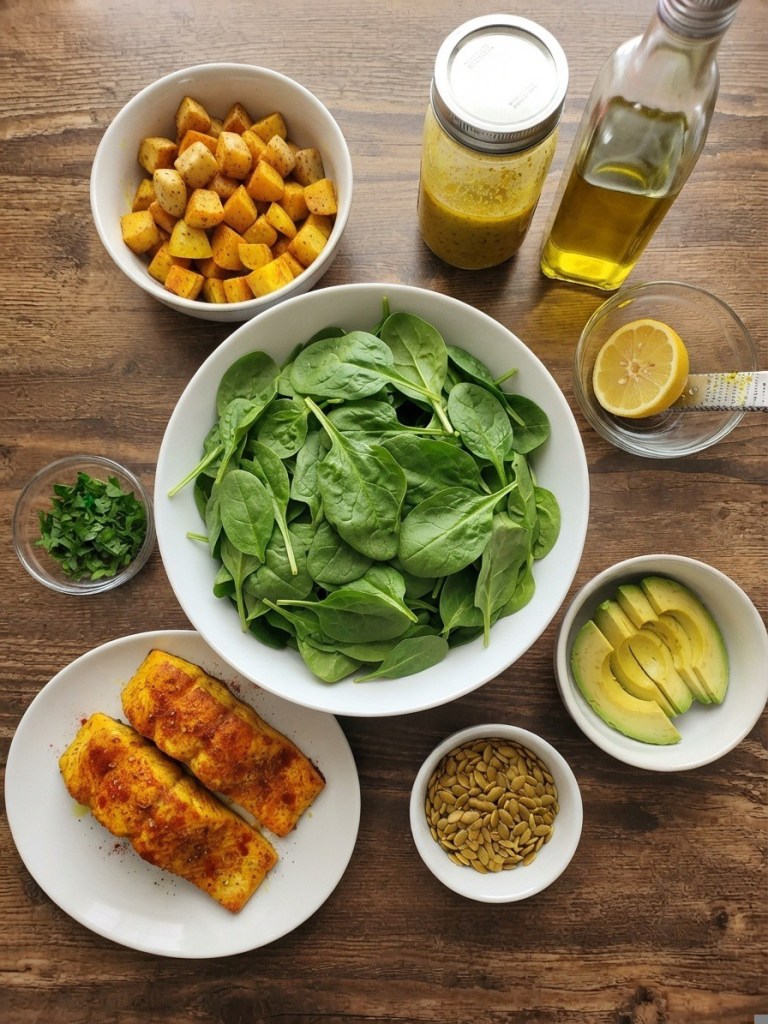 An arrangement of ingredients on a wooden table, including roasted diced potatoes, fresh spinach leaves, sliced avocado, pumpkin seeds, lemon wedges, a glass of olive oil, and a jar of salad dressing, with two pieces of seasoned fish on a plate.