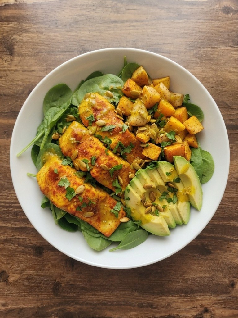 A bowl of spinach topped with grilled fish fillets, roasted squash cubes, sliced avocado, and sprinkled with pumpkin seeds and fresh herbs.