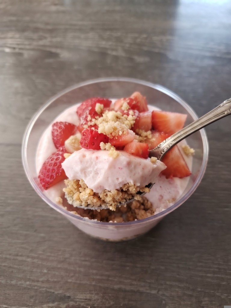 A close-up view of a parfait in a clear cup, featuring layers of creamy strawberry yogurt, topped with fresh strawberry pieces and crumbled granola, with a spoon resting against the edge.
