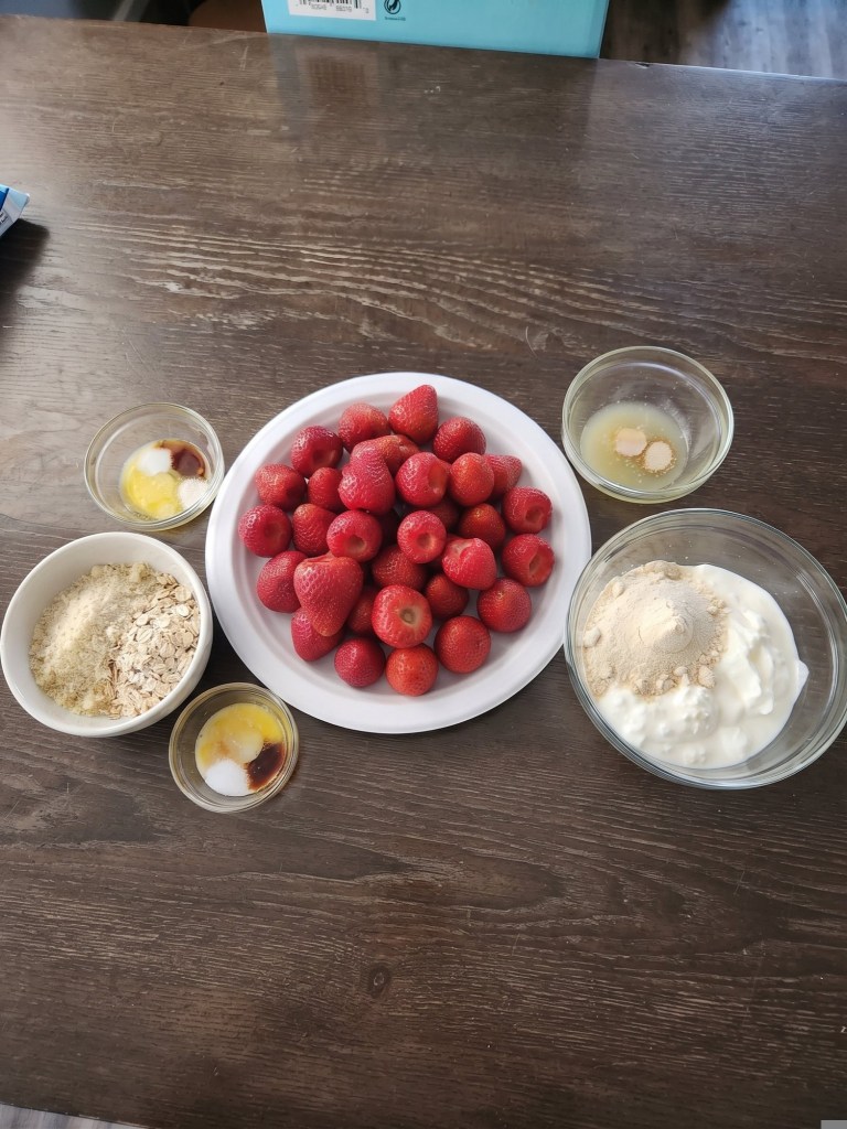 A plate of fresh strawberries surrounded by various ingredients including yogurt, oats, and small bowls with honey and vanilla extract on a wooden table.