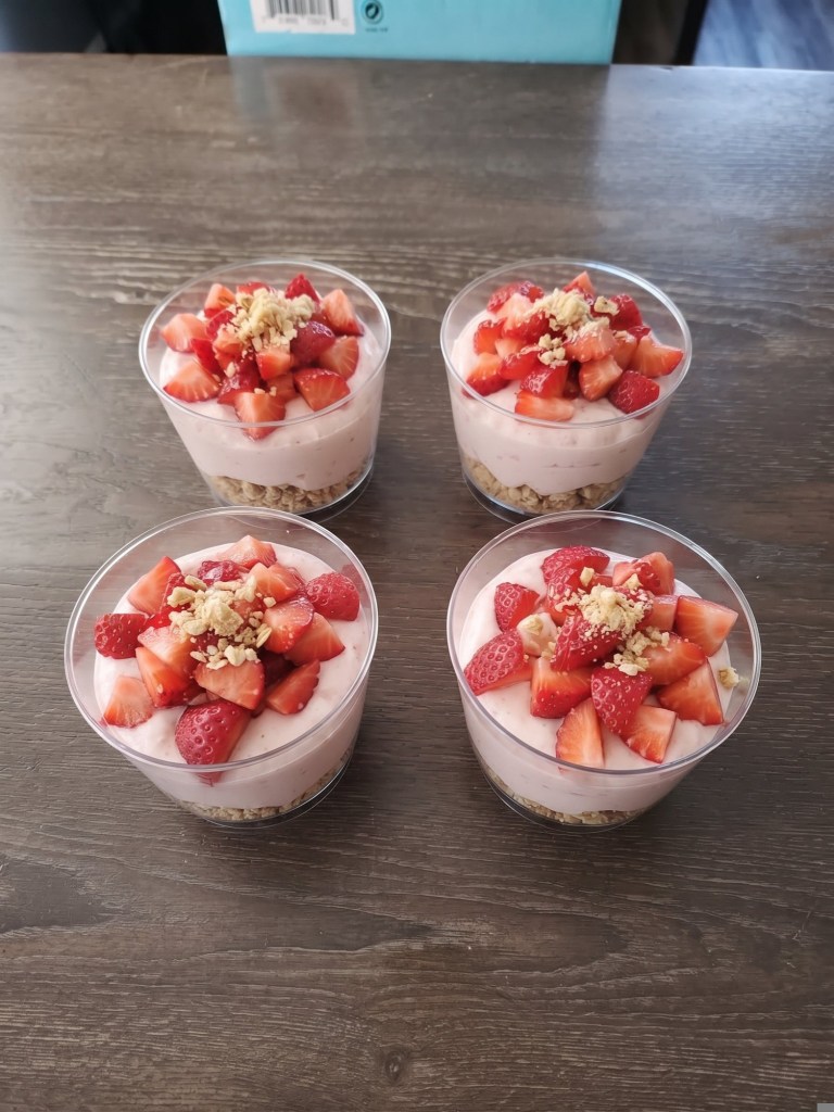 Four cups of layered yogurt desserts topped with fresh strawberries and granola, placed on a wooden surface.