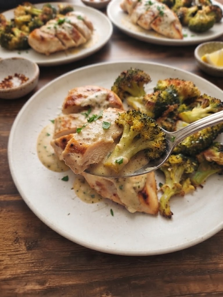 A plate with sliced chicken breast drizzled with sauce, alongside roasted broccoli. A fork is lifting a piece of chicken with broccoli on a wooden table.