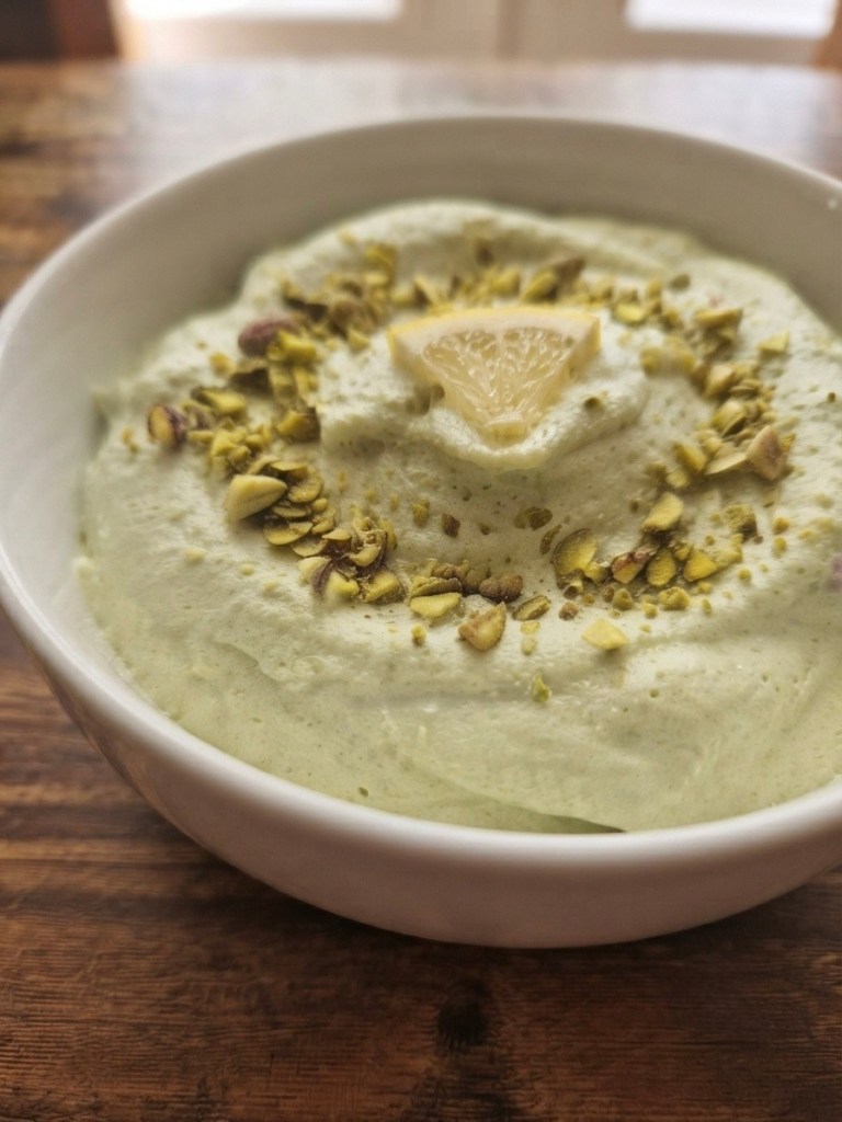A bowl of green dessert topped with crushed pistachios and a lemon slice, placed on a wooden surface.