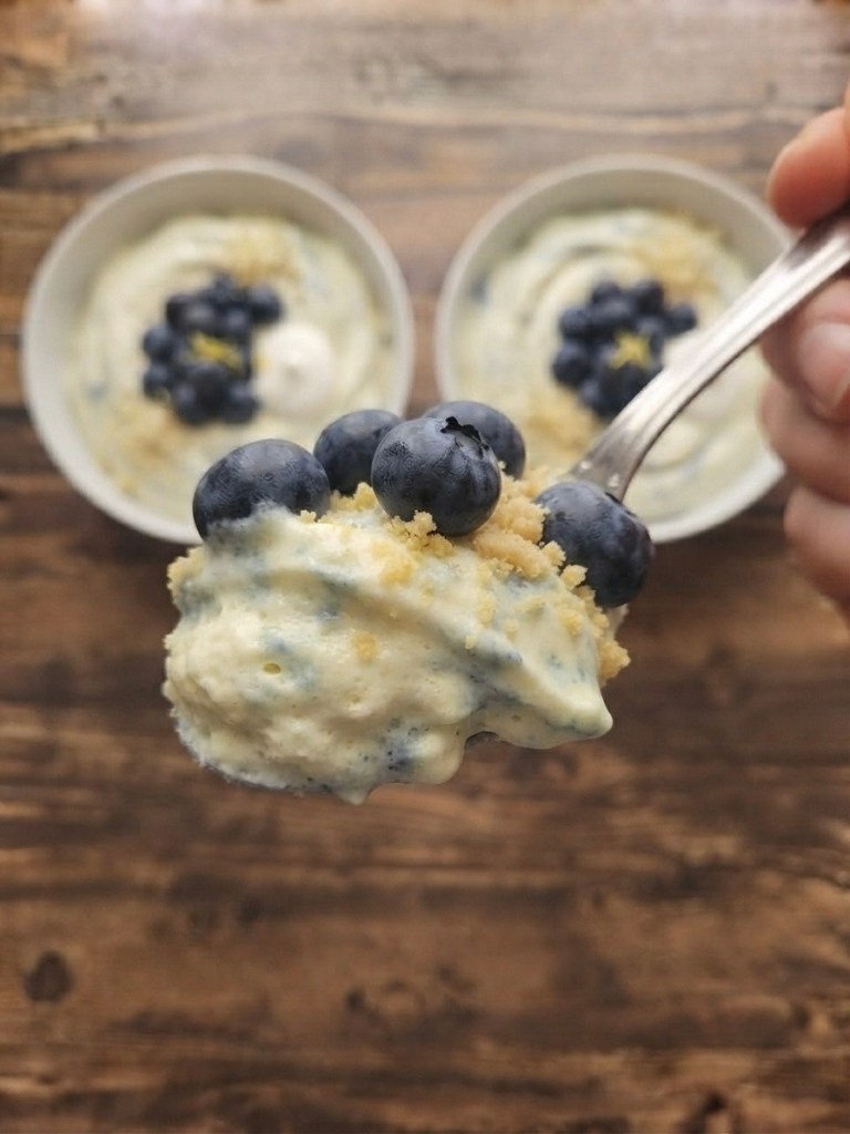 A close-up of a spoonful of dessert topped with blueberries, with two bowls of the same dessert in the background, resting on a wooden table.