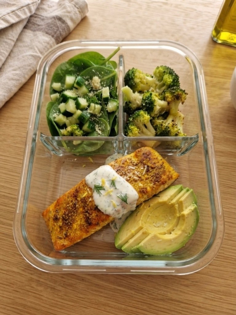 A healthy meal prep container with grilled salmon topped with dill sauce, avocado slices, a spinach and cucumber salad, and steamed broccoli.