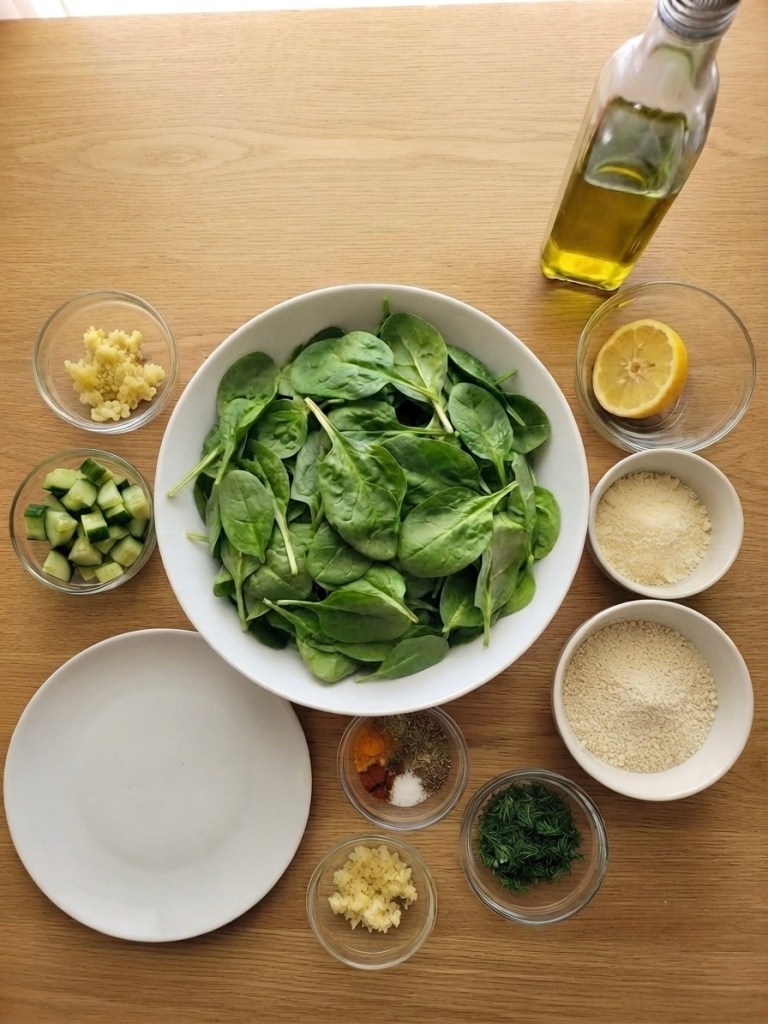 A variety of fresh ingredients for a salad, including a large bowl of spinach, diced cucumber, minced garlic, a sliced lemon, olive oil, and various spices in small bowls, arranged on a wooden table.