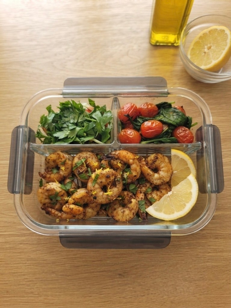 Meal prep container filled with seasoned shrimp, fresh parsley, cherry tomatoes, and spinach, garnished with lemon slices.