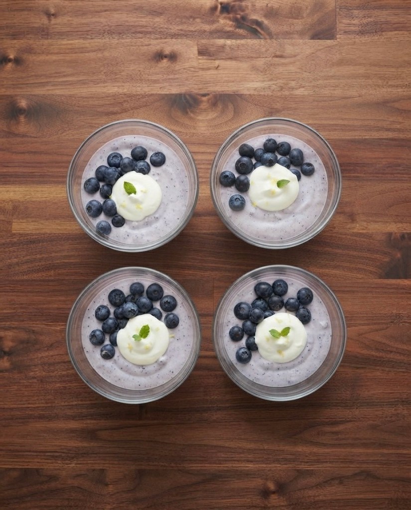 Four glass bowls of blueberry yogurt topped with fresh blueberries and a dollop of whipped cream, arranged on a wooden surface.