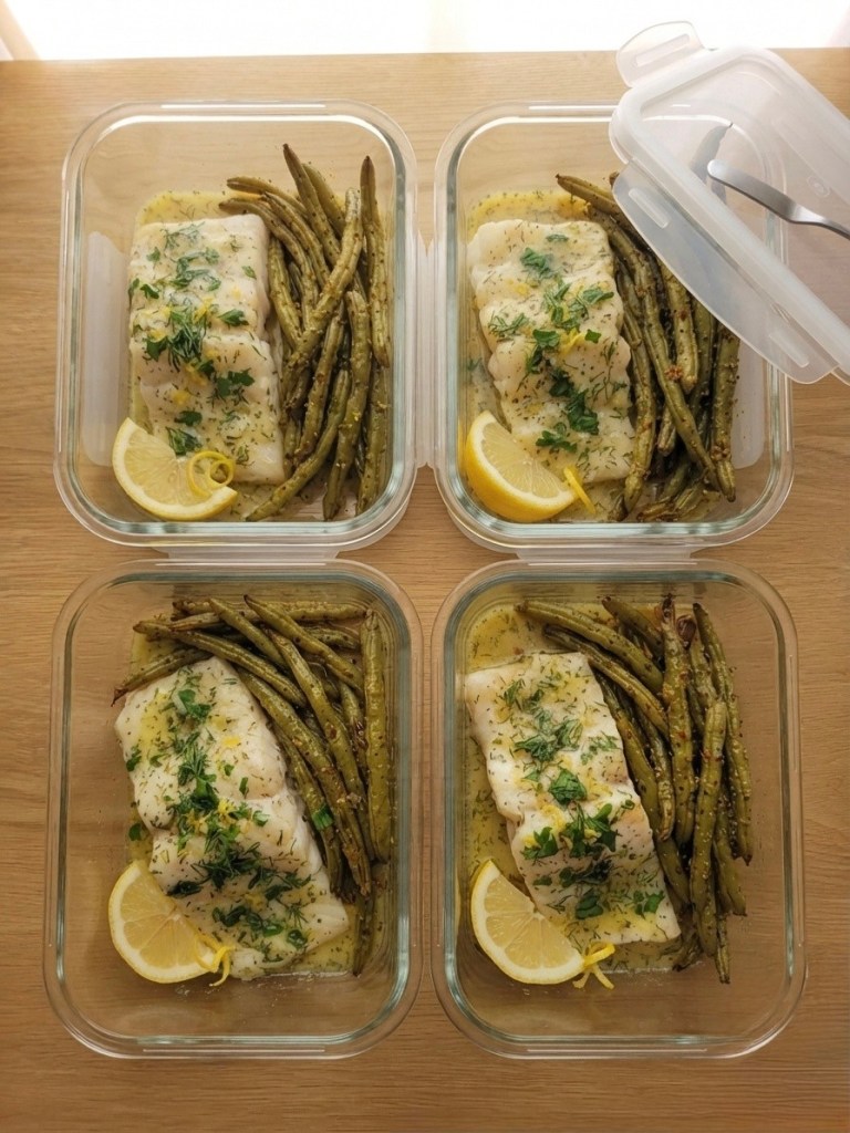Four glass meal prep containers filled with cooked fish and green beans, garnished with lemon slices and herbs.