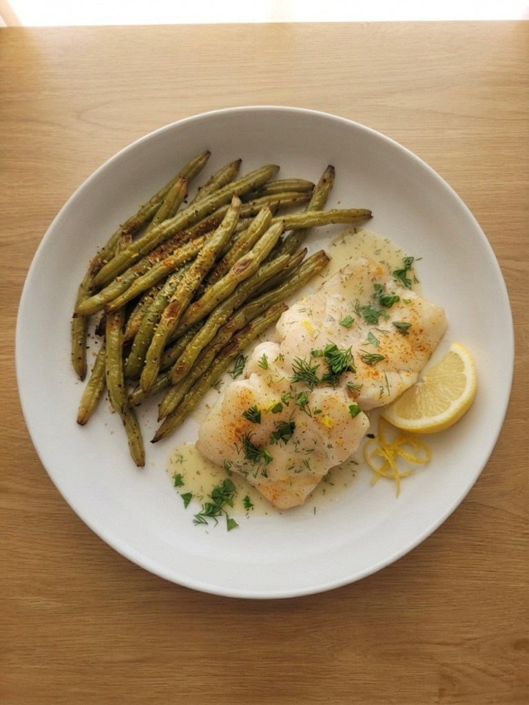 Plate of cooked fish fillets topped with herbs and accompanied by roasted green beans and a slice of lemon.