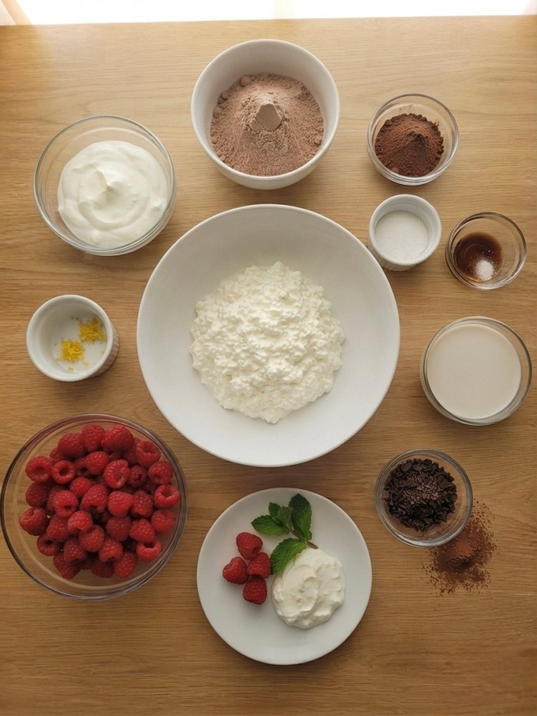 A variety of ingredients for a dessert arranged on a wooden surface. Includes bowls of cottage cheese, yogurt, cocoa powder, and sugar, along with fresh raspberries, mint leaves, and chocolate sprinkles.
