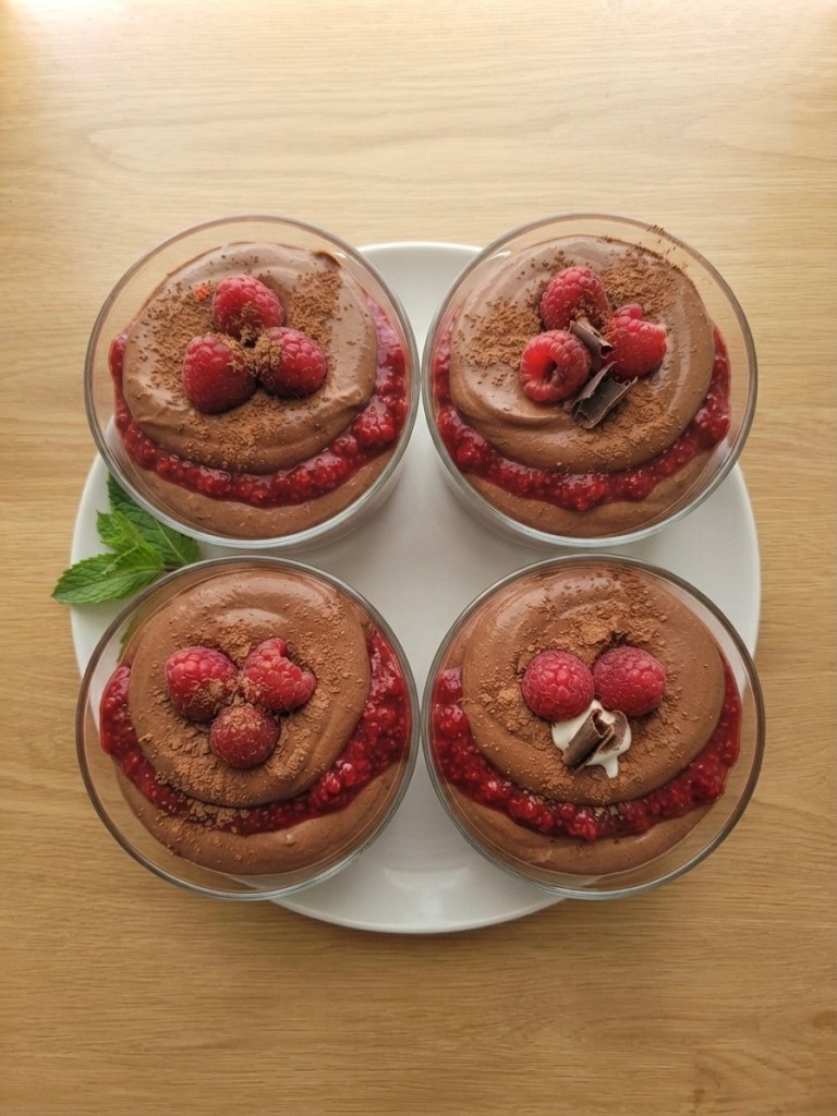 Four dessert cups with chocolate mousse topped with raspberries and cocoa powder, served on a white plate with a mint leaf.