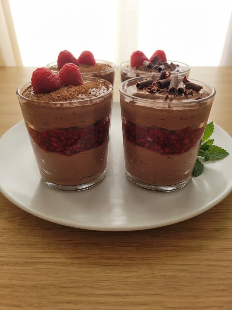 Four dessert glasses containing chocolate mousse with raspberry layers, topped with fresh raspberries and chocolate shavings, on a white plate.
