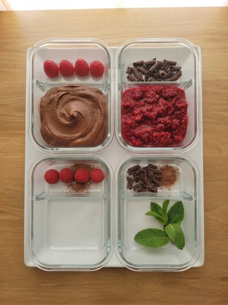 Four glass containers arranged on a wooden surface, containing chocolate mousse, fresh raspberries, raspberry sauce, chocolate shavings, and a sprig of mint.
