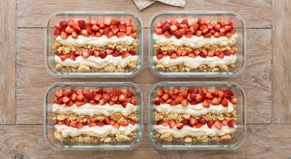 Four glass containers filled with layers of crushed cookies, creamy filling, and topped with fresh strawberries, arranged on a wooden table.